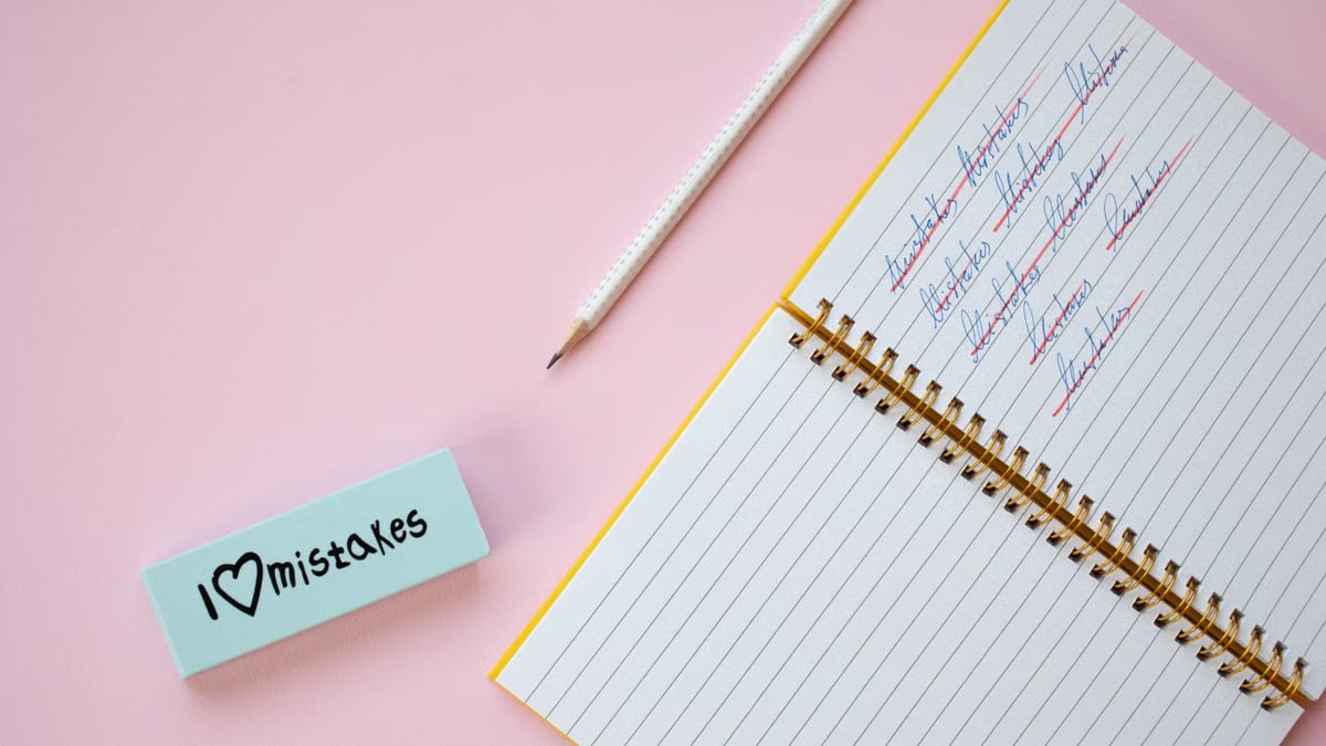Flat lay of a spiral notebook and eraser on a pastel pink background with crossed out words.